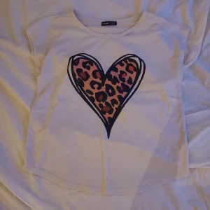 Vit t-shirt med leopardhjärta från Shein - Säljer en vit t-shirt från Shein med ett stort hjärta i leopardmönster på framsidan. T-shirten har en rund halsringning och korta ärmar. Perfekt för en avslappnad stil.