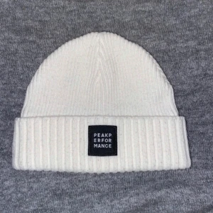 Vit ribbad mössa från Peak Performance - Snygg vit ribbad mössa från Peak Performance. Perfekt för kyliga dagar med stilren design och uppvikt kant. Märkeslogga framtill ger en extra touch.