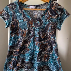 Mönstrad kortärmad topp - Säljer en färgglad topp från Monki med ett livligt paisleymönster i blått, orange och svart. Toppen har en v-ringad hals och korta ärmar, perfekt för en avslappnad stil.