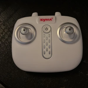 Vit fjärrkontroll och drönare från Syma - Säljer en vit fjärrkontroll och en svart drönare från Syma. Fjärrkontrollen har två styrspakar och en strömknapp i mitten. Drönaren har fyra vita propellrar och en kompakt design. Perfekt för nybörjare som vill utforska drönarflygning!