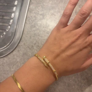 Guldarmband med glittriga detaljer - Elegant guldarmband med en unik design som liknar en spik, dekorerat med glittrande stenar. Perfekt för att ge en lyxig touch till din stil. Armbandet har en öppen design som gör det enkelt att justera. Aldrig använt 