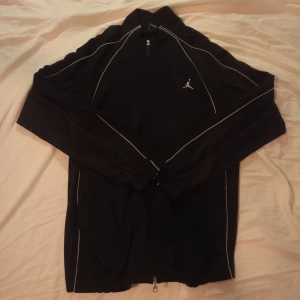 Jordan zip tröja - Snygg svart träningsjacka från Jordan med vita detaljer och dragkedja. Perfekt för sportiga aktiviteter eller som en stilren vardagsjacka. Den har långa ärmar och en liten logga på bröstet.