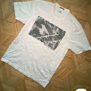 Vit t-shirt från C.P. Company - Säljer en vit t-shirt från C.P. Company med ett tryck från 'The Metropolis Series' på framsidan. T-shirten har korta ärmar och är tillverkad i ett mjukt material. Perfekt för en avslappnad stil.  Priset kan sänkas vid snabb affär.
