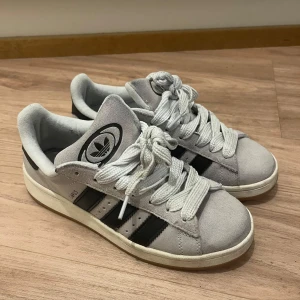 Grå Adidas Campus sneakers - Snygga grå Adidas Campus sneakers (storlel 40.5) i mocka med klassiska svarta ränder. Skorna har en vit sula och breda snören för en stilren look. Perfekta för en avslappnad stil.