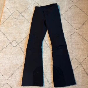 Svarta täckbyxor från Peak Performance - Snygga svarta täckbyxor från Peak Performance med dragkedjor vid bensluten och broderad logga. Den har RECCO advanced rescue teknologi. Byxorna har även snölås. Perfekta för slalom med slitstarkt material och praktiska detaljer.