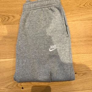 Grå mjukisbyxor från Nike - Säljer ett par grå mjukisbyxor från Nike med elastisk midja och sidofickor. Byxorna har en avslappnad passform och är perfekta för en bekväm dag. Nike-loggan är broderad i vitt på benet.