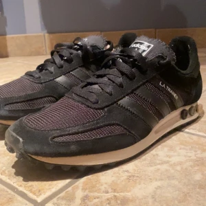 Svarta Adidas L.A. Trainer sneakers - Snygga svarta Adidas L.A. Trainer sneakers med klassiska tre ränder i svart och vit sula. Skorna har snörning och en retro design med dämpande pluggar i sulan. Perfekta för en sportig look.