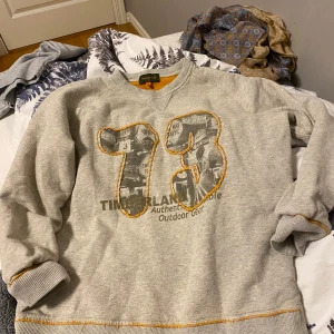 Grå sweatshirt från Timberland - Säljer en grå sweatshirt från Timberland med trycket '73' i svart och orange detaljer. Tröjan har långa ärmar och är perfekt för en avslappnad stil. Den är gjord i mjukt material och har en rund halsringning.