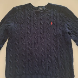 Stickad Ralph Lauren tröja - Säljer en snygg mörkblå stickad tröja från Ralph Lauren med kabelstickat mönster och röd logga på bröstet. Perfekt för kyliga dagar med sin klassiska design och bekväma passform.