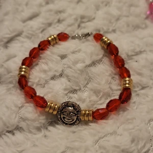 Rött armband med solberlock - Charmigt armband med röda pärlor och guldfärgade detaljer. Armbandet har en unik solberlock i mitten som ger en bohemisk känsla. Perfekt för att lägga till en färgklick till din outfit.