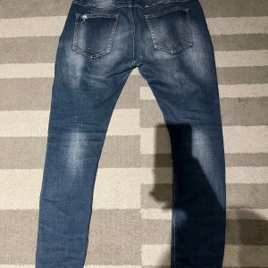 Blå slitna jeans - Säljer ett par blå jeans med slitningar och en cool, distressed look. De har en klassisk femficksdesign och en knappgylf. Perfekta för en avslappnad stil.