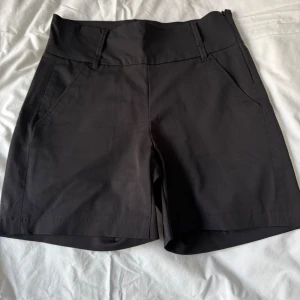 Svarta Fleur shorts från House of Lola - Snygga svarta shorts från House of Lola med dragkedja i sidan och en bred skön linning. Sidofickor och dekorativa bakfickor. Tillverkade i en bekväm mix av bomull, polyamid och elastan för en skön och stretchig passform. Perfekta för en stilren look.