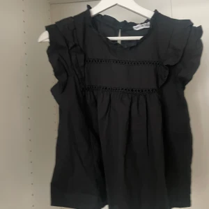 Svart blus från Meet Me There - Snygg svart blus från Meet Me There med volangdetaljer vid ärmarna och spetsdetaljer framtill. Perfekt för en stilren look.