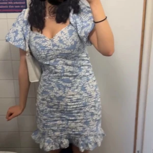 Blommig blå klänning - Charmig blå klänning med vita blommor och volangdetaljer. Den har en åtsittande passform med korta ärmar och en söt volangkant längst ner. Perfekt för somriga dagar!🦋