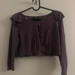 Lila kofta från Soaked in Luxury - Säljer en lila kofta från Soaked in Luxury med volangdetaljer vid axlarna. Koftan är croppad och har trekvartsärmar samt knappar framtill. Perfekt för att lägga till en färgklick i din outfit!
