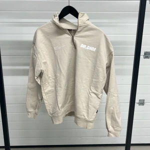 Beige tröja med dragkedja från Dr. Denim - Säljer en stilren beige tröja från Dr. Denim med halv dragkedja och tryck på bröstet. Tröjan har långa ärmar och ribbade muddar vid ärmslut och nederkant. Perfekt för en avslappnad look.