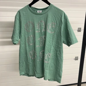 Grön t-shirt med strassdetaljer från One of One - Snygg grön t-shirt från One of One med glittrande strassdetaljer på framsidan. Perfekt för att ge din outfit lite extra bling. T-shirten har en normal passform och korta ärmar.
