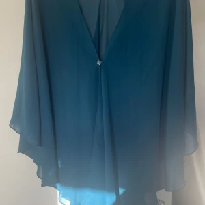 Blå chiffongblus - Elegant blå chiffongblus med v-ringad design och en dekorativ knapp framtill. Blusen har en luftig passform och långa, fladdriga ärmar som ger en stilfull look.