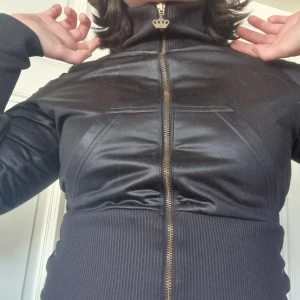 Svart jacka med gulddetaljer från Adidas - Snygg svart jacka från Adidas med gulddragkedja och knappar, äkta 2000tal.. Jackan har en cool krona i strass på ryggen och ribbade muddar. Perfekt för en stilren look.