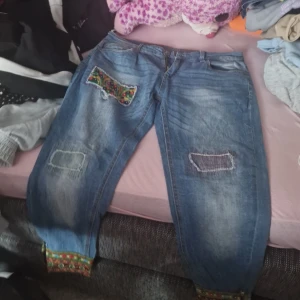 Unika jeans med broderade detaljer - Säljer ett par coola jeans från Desigual i storlek 30. De är blå med unika broderade detaljer i färgglada mönster på fickorna. Jeansen har en sliten look med patchar och är perfekta för en avslappnad stil. De har en knapp och dragkedja framtill.