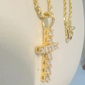 Guldhalsband med korshänge - Säljer ett elegant guld färgat halsband med ett korshänge. Hänget är dekorerat med glittrande stenar som ger en lyxig känsla. Perfekt för både vardag och festliga tillfällen. Kedjan är tvinnad och har en säker låsning. NYTT. Ca 50 cm