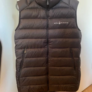 Svart dunväst från Sail Racing - Säljer en svart dunväst från Sail Racing. Västen har en dragkedja framtill och två praktiska fickor med dragkedjor. Perfekt för kyliga dagar när du vill hålla värmen utan att kompromissa med stilen. Den är i ny skick, ny pris 2200.