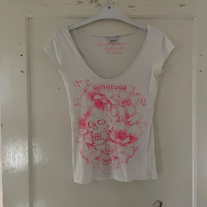Vit t-shirt med rosa tryck från Gina Tricot - Säljer en vit t-shirt från Gina Tricot i storlek XS. Den har ett coolt rosa tryck med texten 'Gorgeous' och 'Rock' samt blommor och en dödskalle. T-shirten har en djup v-ringning och korta ärmar. Perfekt för en avslappnad och rockig stil!