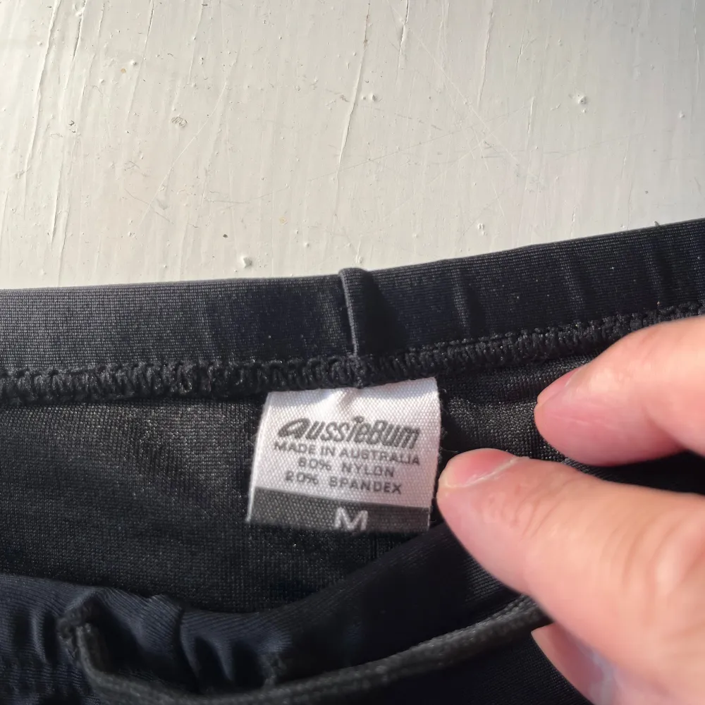 Säljer ett par snygga svarta badshorts från AussieBum med texten 'Australia' i gult och blått på framsidan. De är gjorda av 80% nylon och 20% spandex, vilket gör dem superbekväma och perfekta för en dag på stranden. Shortsen har en tight passform och en liten röd etikett på sidan. Perfekta för sommarens alla badäventyr! 🏖️. Shorts.