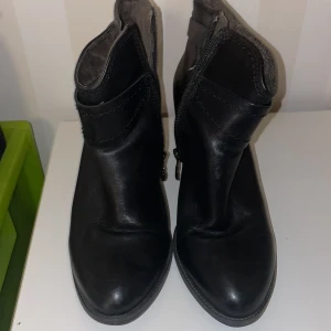Tamaris boots med klack och dragkedja - Snygga svarta boots från Tamaris med klack och dekorativa spännen knappt använda. Skorna har en praktisk dragkedja på sidan för enkel på- och avtagning. Perfekta för en stilren look.