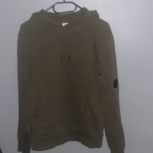 Olivgrön hoodie från C.P. Company - Säljer en olivgrön hoodie från C.P. Company med en unik detalj på ärmen. Perfekt för en avslappnad stil. Hoodien har en klassisk design med en bekväm passform. Skick 8/10 