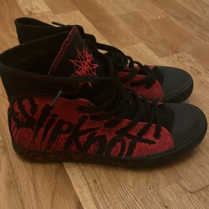 Svarta och röda sneakers med Slipknot-design - Unika sneakers i svart och rött med Slipknot-logga och detaljer. Skorna har hög snörning och en cool design med bandets ikoniska symboler. Perfekta för fans av bandet som vill ha en edgy stil.  Skorna har bara varit använda en gång. Originella skokartonger tillkommer. Storlek 41. Köpare står för frakt