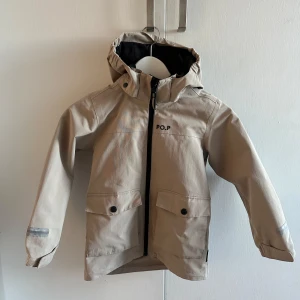 Beige vindjacka från Polarn O. Pyret - Säljer en beige vindjacka från Polarn O. Pyret med huva och dragkedja. Jackan har långa ärmar och praktiska fickor framtill. Perfekt för blåsiga dagar.