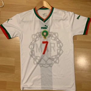 Morocco 🇲🇦 borta 2022/23 Ziyech - Säljer en vit fotbollströja från Puma med gröna och röda detaljer. Tröjan har korta ärmar och nummer 7 på framsidan samt 'ZIYECH' på baksidan. Väldigt bra skick med starkt tryck och fina färger därav priset.