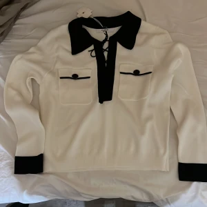 Vit tröja med svarta detaljer från ELSA.M - Snygg vit tröja från ELSA.M med svarta detaljer. Tröjan har en krage och snörning framtill samt två dekorativa fickor med knappar. Perfekt för en stilren look.