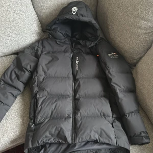 Svart dunjacka från Sail Racing. Nytt skick passar perfekt för vintern! - Säljer en svart dunjacka från Sail Racing med huva och dragkedja. Jackan har en broderad logga på ärmen och är perfekt för kalla dagar. Designad för att klara extrema väderförhållanden.