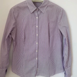 Randig skjorta med knappar - Snygg randig skjorta i vitt och lila med klassisk krage och knappar framtill. Perfekt för en stilren look.                                                                     Skuldra - Skuldra 42 cm.                                    Längd från axel 61                                                Längd från armhåla 37.                                         Över byst 49 cm