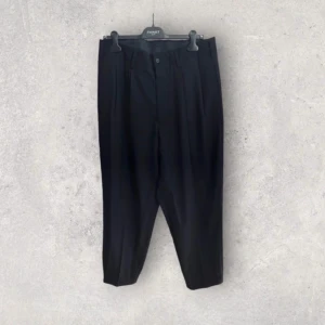 Yohji Yamamoto Svarta kostymbyxor - Snygga svarta kostymbyxor från Yohji Yamamoto med hög midja och rak och lös passform. Byxorna har knäppning fram och är perfekta för en stilren look. Passar utmärkt till både vardag och mer formella tillfällen.