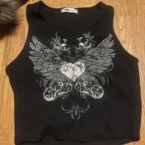 Svart ribbad linnetopp med tryck - Snygg svart ribbad linnetopp från Sister med ett coolt tryck av vingar och hjärta i grått och vitt. Perfekt för en edgy look!