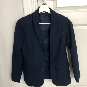 Mörkblå kavaj från Lindex - Snygg mörkblå kavaj från Lindex för barn i storlek 152. Kavajen har en klassisk design med knappar framtill och långa ärmar. Perfekt för både vardag och speciella tillfällen.