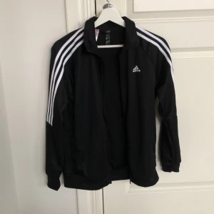 Svart träningsjacka från Adidas - Säljer en svart träningsjacka från Adidas med de klassiska tre vita ränderna längs ärmarna. Jackan har dragkedja och är långärmad, perfekt för sportiga aktiviteter. Tillverkad av Primegreen-material.