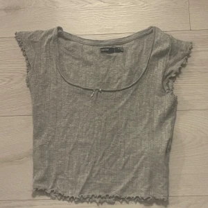 Grå ribbad topp från House - Säljer en söt grå ribbad topp från House i storlek XS. Toppen har korta ärmar och en liten rosett framtill. Perfekt för en avslappnad stil.