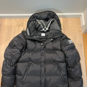 Svart dunjacka från Burberry - Säljer en stilren svart dunjacka från Burberry med avtagbara ärmar och klassiskt rutigt foder i huvan. Jackan har dragkedja och knappar framtill samt en logotyp på ärmen. Perfekt för kyliga dagar.