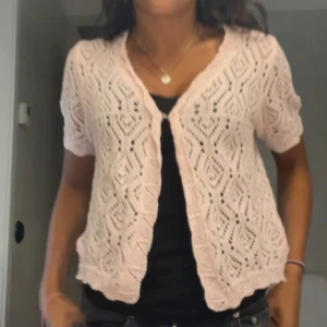 Rosa stickad kofta - Söt rosa stickad kofta med kortärmad design och ett fint hålmönster. Perfekt för lager-på-lager-stil och ger en romantisk touch till din outfit.