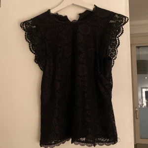 Svart spetsblus från Pieces - Snygg svart blus från Pieces med vackra spetsdetaljer. Perfekt för en elegant look. Blusen har korta ärmar och en rund halsringning.