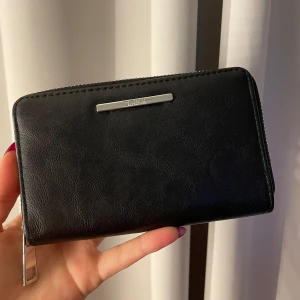 Svart plånbok med dragkedja - Snygg svart plånbok i skinn med dragkedja runtom. Den har flera fack för kort och sedlar, samt en stilren metallplatta på framsidan. Perfekt för att hålla ordning på dina värdesaker.