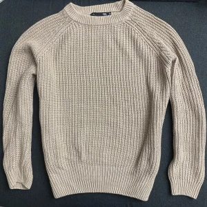 Beige stickad tröja - Säljer en stilren beige stickad tröja med rund hals och långa ärmar. Perfekt för kyligare dagar och passar till det mesta i garderoben. Tröjan har en klassisk design med ribbstickade muddar vid ärmslut och nederkant.