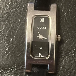 Svart Gucci klocka - Elegant rektangulär klocka från Gucci med svart urtavla och romerska siffror. Klockan har en stilren design med ett svart skinnarmband och silverfärgad boett. Perfekt för en sofistikerad look.