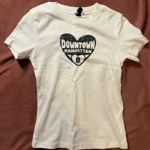 Vit t-shirt med tryck från H&M Divided - Säljer en vit t-shirt från H&M Divided med ett hjärtformat tryck där det står 'Downtown Manhattan' och en liten dödskalle. T-shirten är kortärmad och gjord i bomull, perfekt för en avslappnad stil.