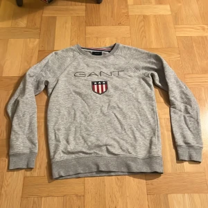 Grå sweatshirt från GANT - Säljer en stilren grå sweatshirt från GANT med broderad logga och sköld på bröstet. Tröjan har långa ärmar och ribbade muddar vid ärmslut och nederkant. 