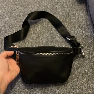 Svart midjeväska med dragkedja - Snygg och praktisk svart midjeväska i syntetmaterial. Väskan har en justerbar rem och en dragkedja för säker förvaring. Perfekt för att hålla dina saker nära till hands.